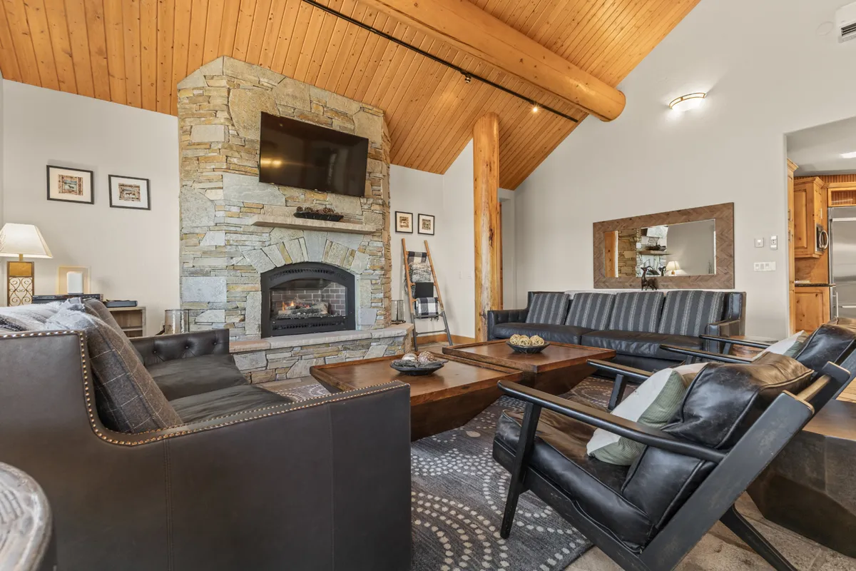 A closer look at the upper living room, this image highlights the towering stone fireplace, massive log beams supporting a vaulted ceiling, plush seating, and a large TV—perfect for relaxing, socializing, or movie nights. The room radiates warmth and comfort, emphasizing the heart-of-the-home atmosphere.
