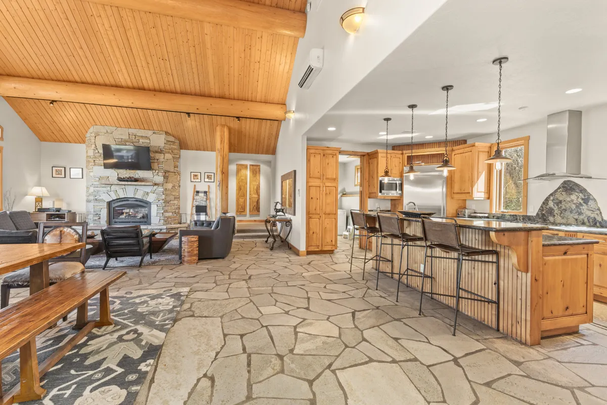A stylish open-concept interior featuring a stone fireplace, kitchen island, wooden beams, and slate flooring. The space is shown as lively and communal, ideal for cooking, entertaining, and relaxing after outdoor adventures.