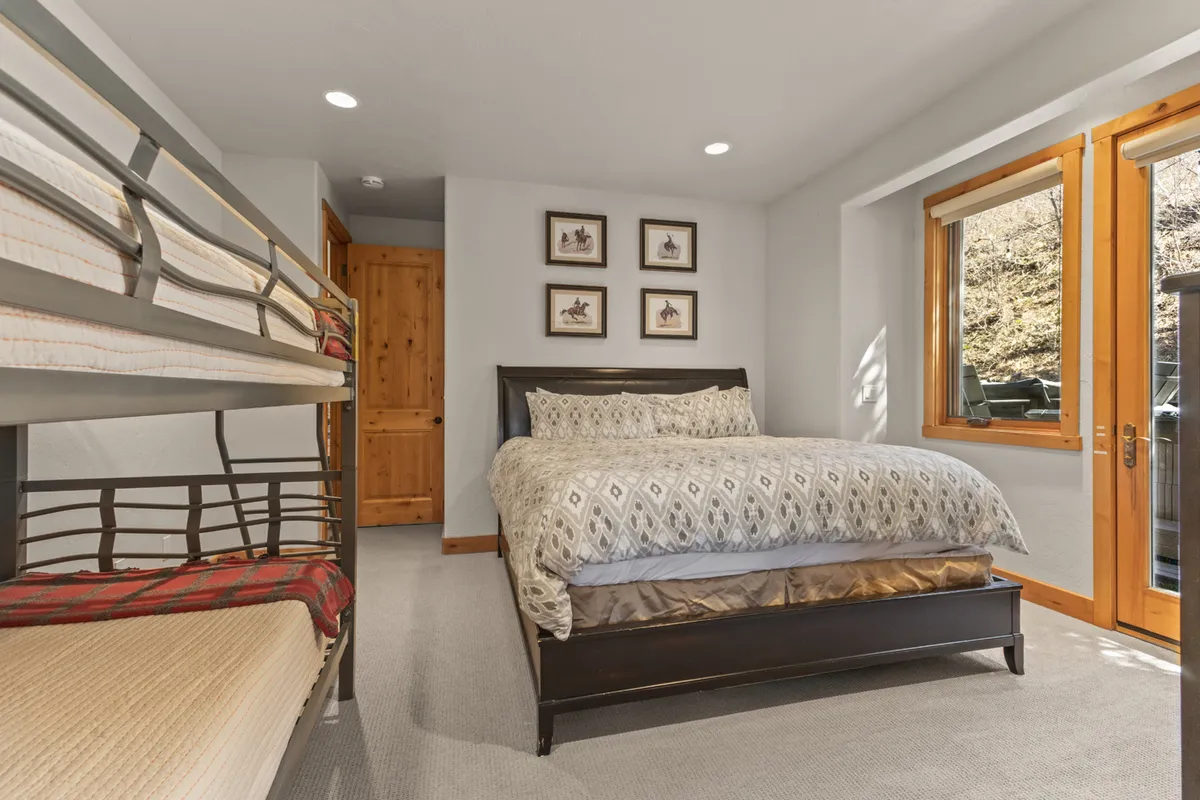 A serene bedroom featuring a queen bed and an adjacent twin bunk bed, framed art on the wall, and ample natural light streaming through large windows. The room is styled for families, with inviting textures and a relaxed, flexible layout.