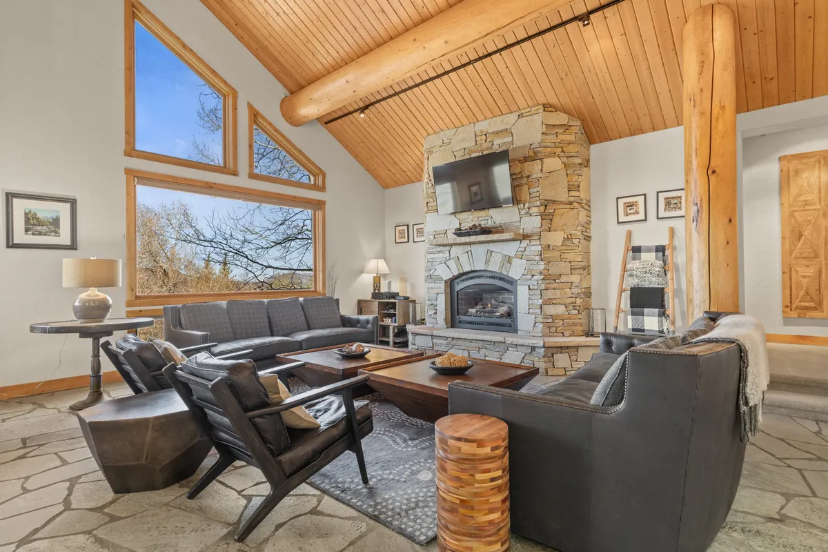 An expansive great room with impressive stone fireplace, tall wooden beams, large windows framing inspiring mountain views, and plush, comfortable seating arranged for conversation and connection.