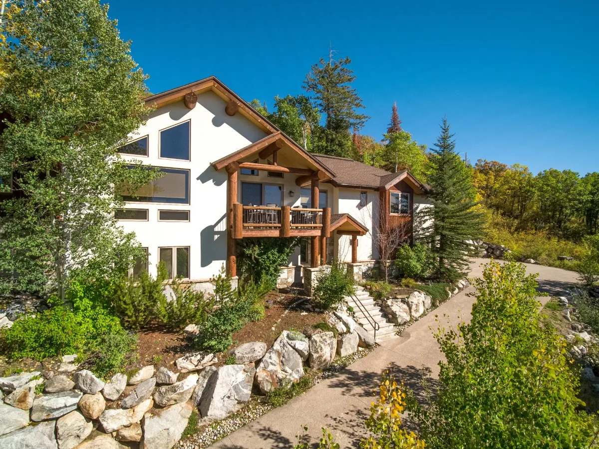 The hero image showcases the chalet's inviting wooden balcony overlooking sweeping mountain views, with large picture windows framing the outdoors and a charming stone pathway winding through a tranquil garden setting. The space radiates warmth and comfort, ideal for outdoor gatherings, relaxing with a morning coffee, or spotting local wildlife at sunset.