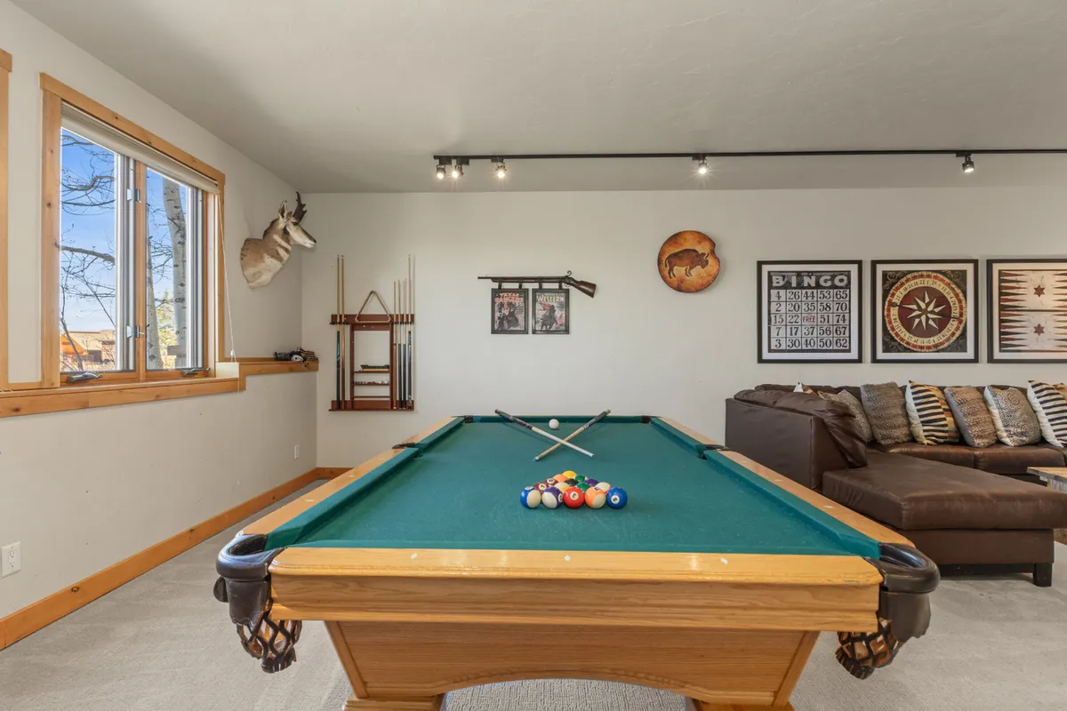 A bright and spacious game room featuring a full-size pool table, a plush sofa, decorative wall art, and a window streaming in natural light. The layout is open and inviting, creating the perfect backdrop for game nights or casual relaxation.