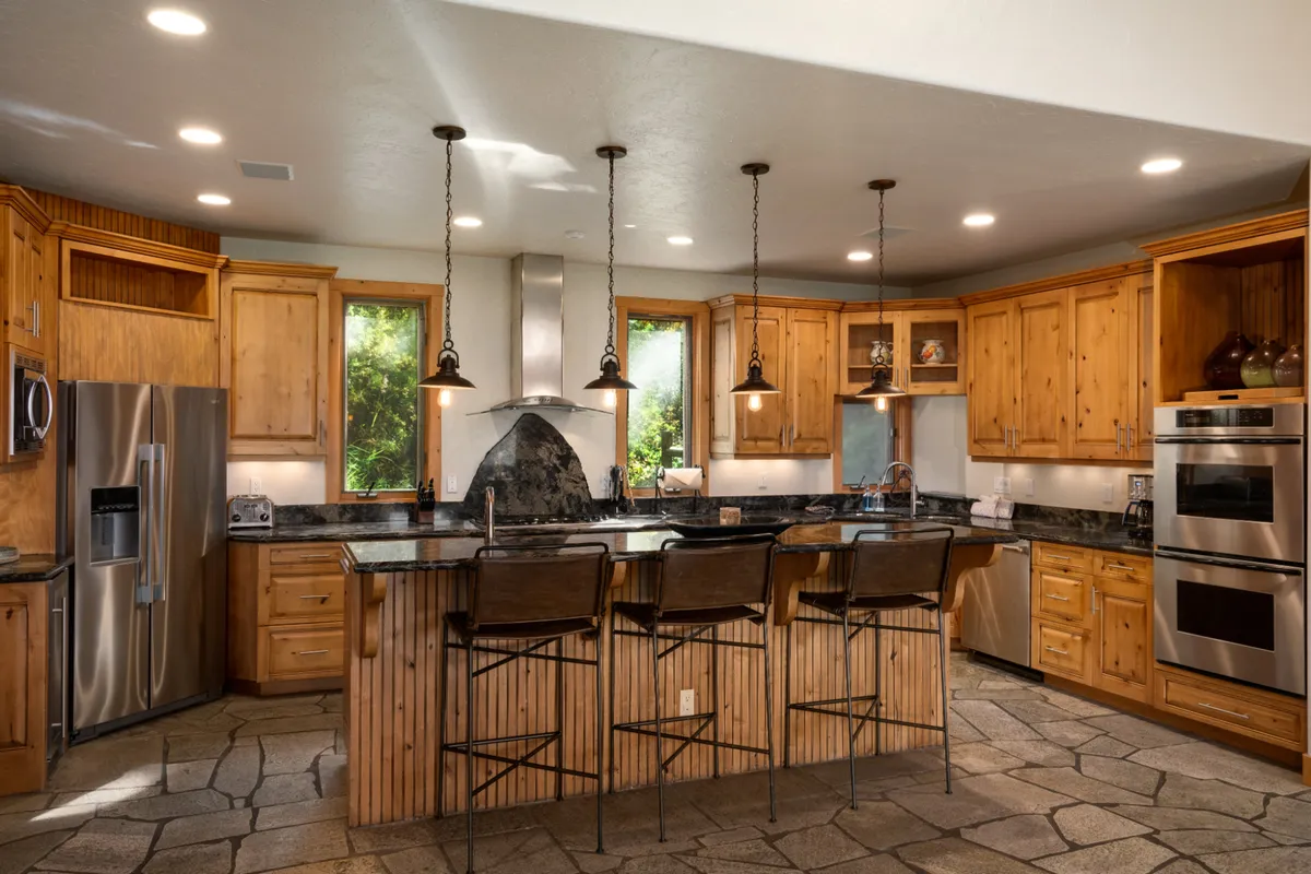 This image centers on the gourmet kitchen’s spacious island with bar stools, surrounded by wooden cabinetry and granite countertops. Ample natural light pours in through large windows, creating a welcoming environment for cooking and casual gatherings. The layout suggests a space designed for both culinary creativity and social interaction.