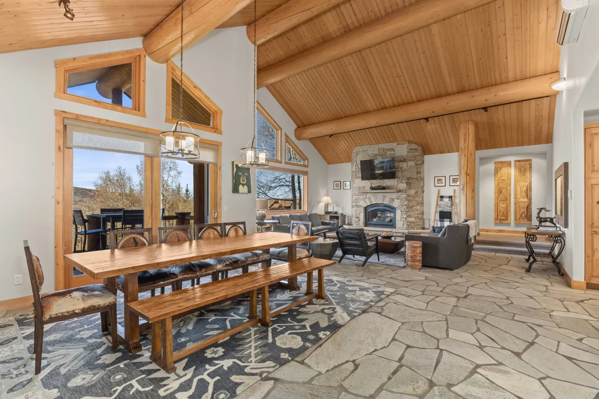 This image showcases a warm, welcoming open-concept space featuring a large wooden dining table set for a group, a substantial stone fireplace, comfortable seating, and expansive picture windows that offer breathtaking views of the mountains and valley beyond. The high wooden beams and natural materials amplify the grandeur and rustic luxury described.