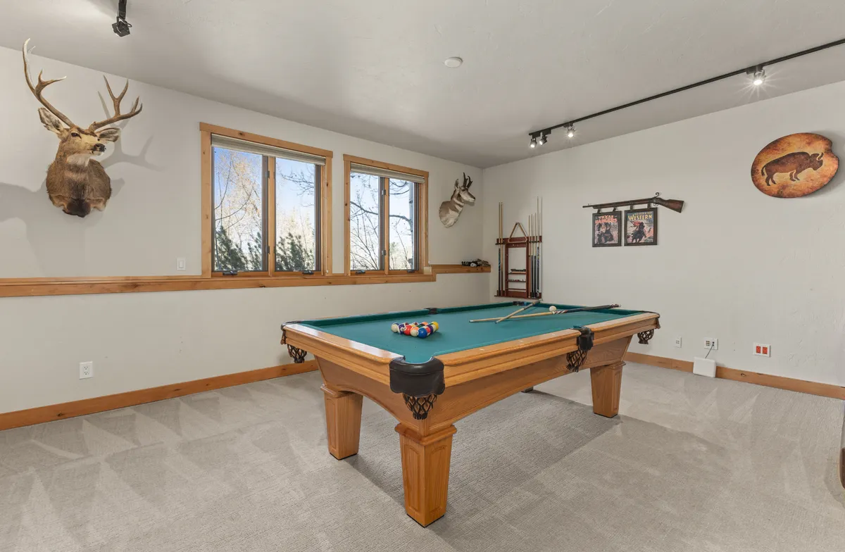 A vibrant interior shot highlighting a pool table in the foreground, natural light pouring through the windows, a mounted deer head for rustic flair, and various pieces of wall art. The overall mood is playful and relaxed, perfectly matching the spirit of a versatile retreat.