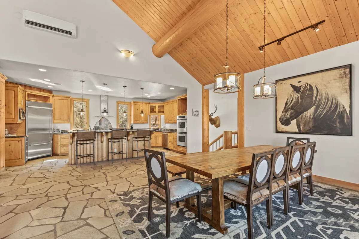 The view extends from the kitchen to a large wooden dining table, suggesting fluidity between food preparation and group dining. The kitchen island is visible, and the space is framed by warm lighting and tasteful artwork. The setting invites celebratory meals and lively gatherings.