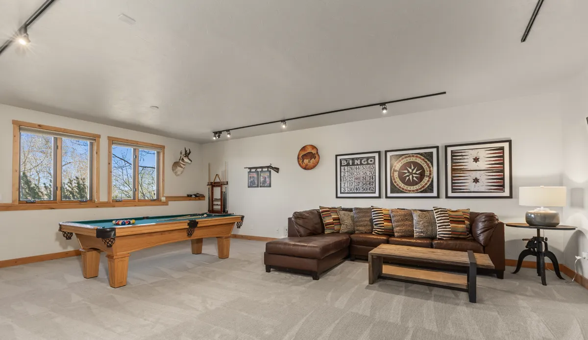 A spacious game room featuring a pool table, a cozy sofa, contemporary artwork, and large windows that flood the space with natural light. The windows hint at the surrounding landscape, creating an inviting environment for both play and relaxation.